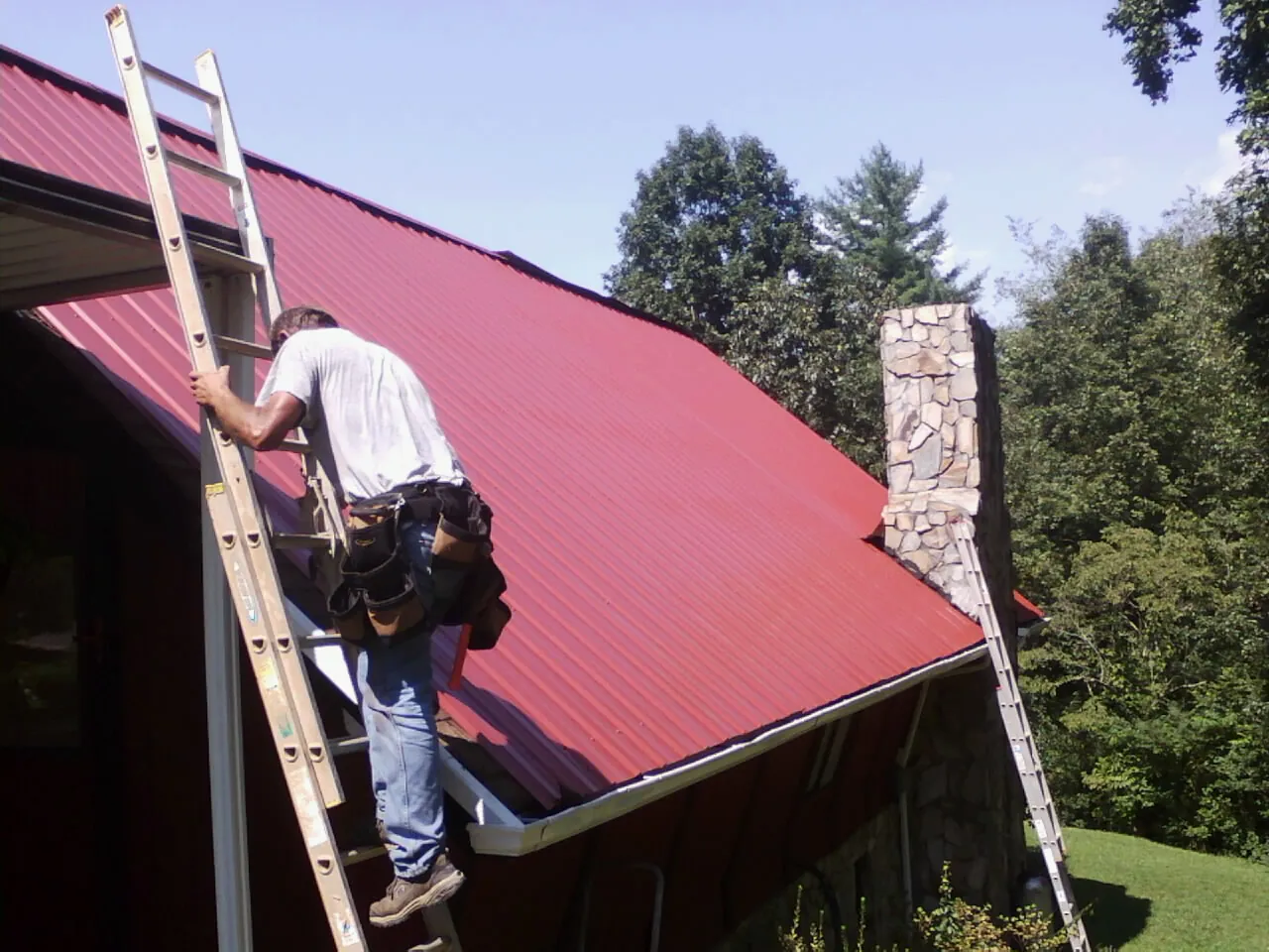 Roofer on a ladder beside a red metal roof and stone chimney — Soffit & Fascia Repair by Roofing Brothers in Check